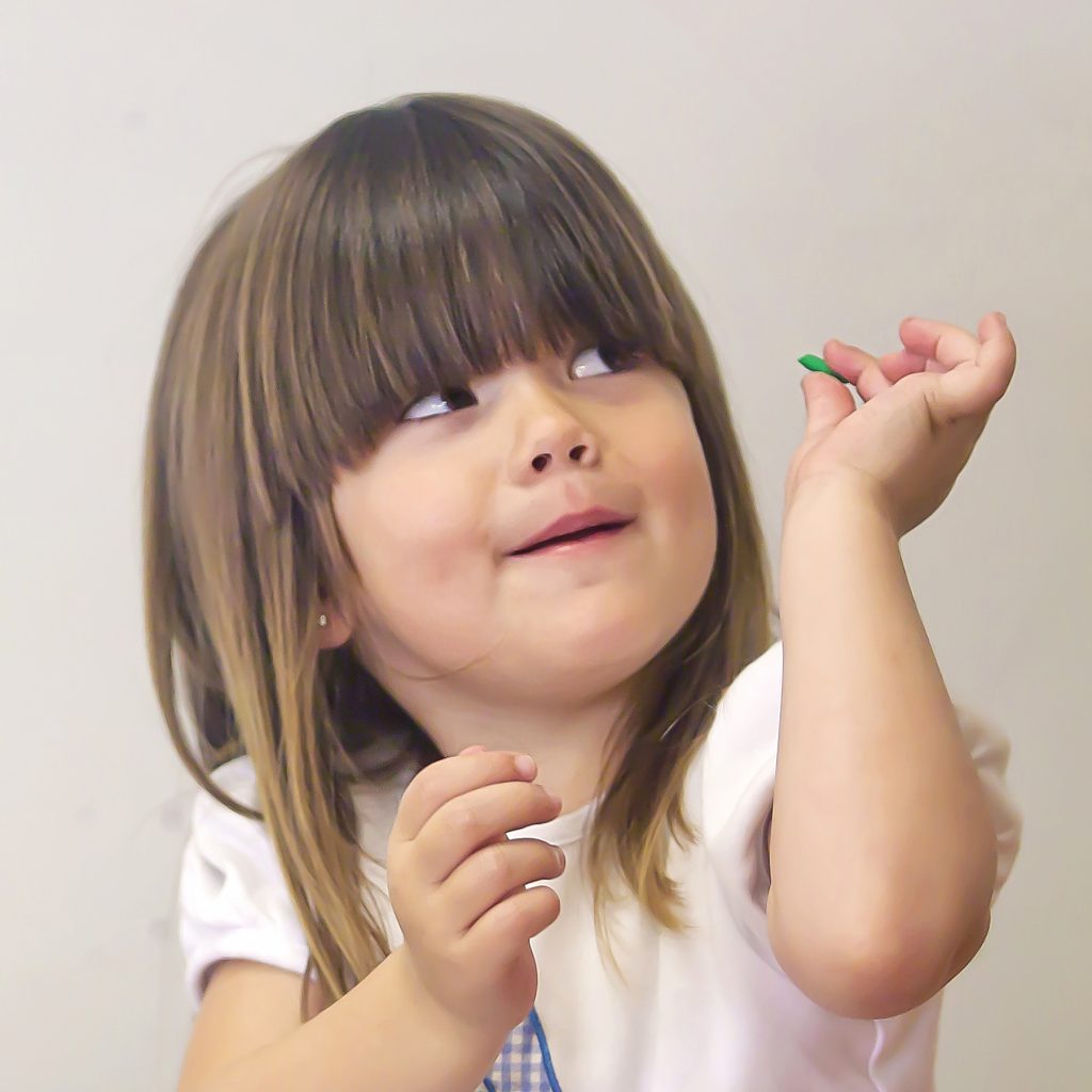 A three year old kindergarten student with brown hair looks up with a happy expression while holding a game piece that helps develop fine motor skills.
